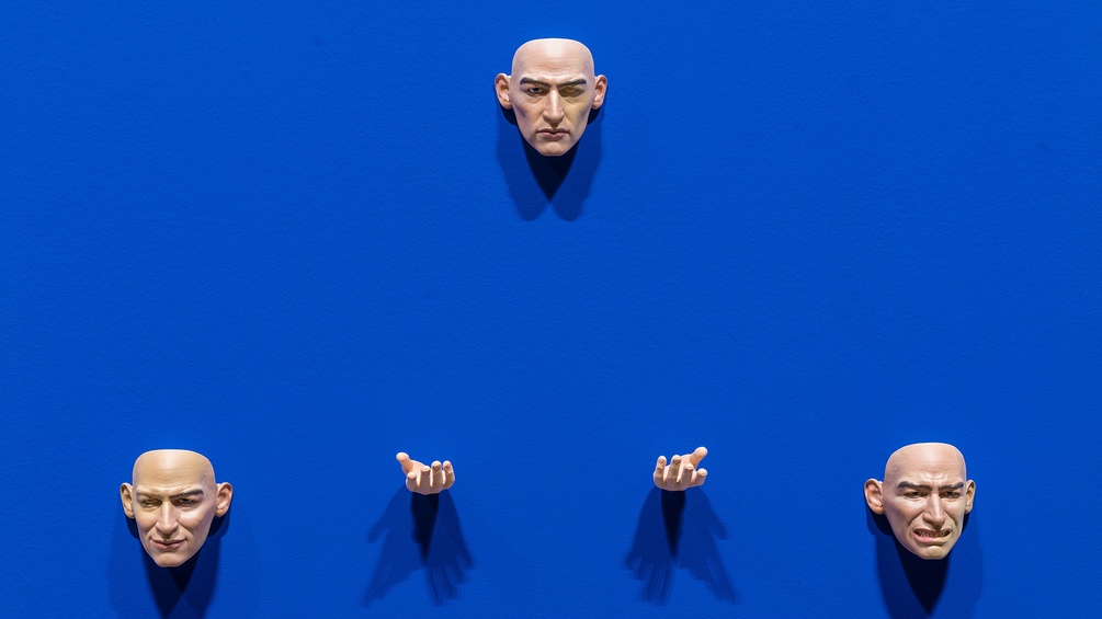 Ausstellungsansicht: Drei Gesichter und zwei Hände auf blauem Hintergrund