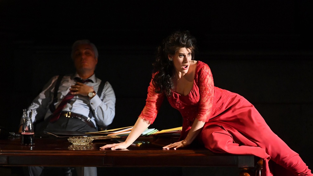 Anja Harteros im roten Kleid auf einem STisch sitzend, dahinter ein Mann
