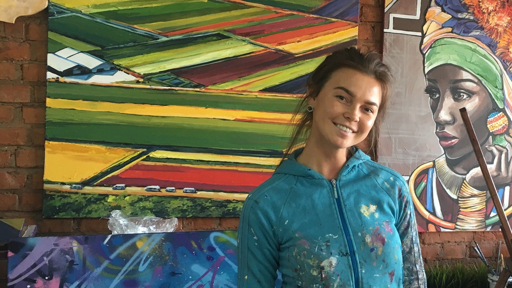 Eine junge Frau steht in ihrem Atelier vor ihren Bildern, ihre Jacke ist voller Farbspritzer.