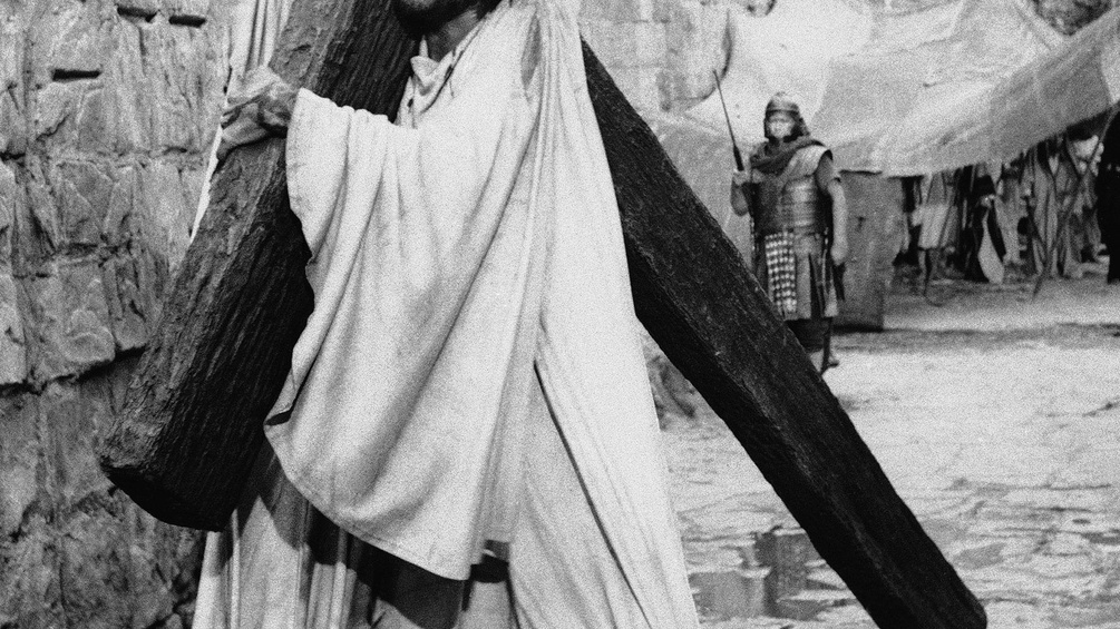 Mann als Jesus verkleidet trägt ein Kreuz, dahinter ein Römer