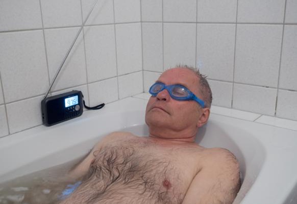 Ein Mann liegt in der Badewanne. Er hat Schwimmbrillen auf. Am Wannenrand ein Radiogerät.