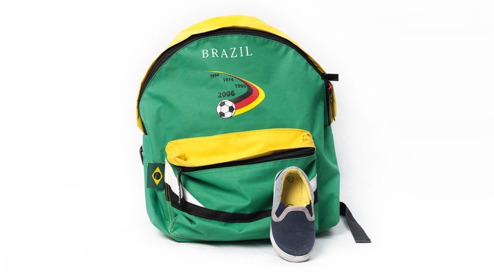 Grün-gelber Rucksack mit der Aufschrift "Brazil" und einem Fußball. Davor ein kleiner dunkelblauer Hausschuh.