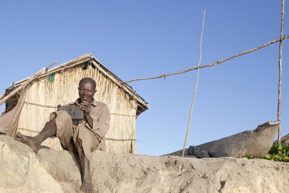 Afrikanischer Mann sitzt mit Radio in der Hand vor einer Stroh-Hütte.