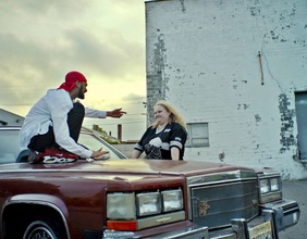 Rapper auf Motorhaube zeigt auf Frau mit blonden Haaren