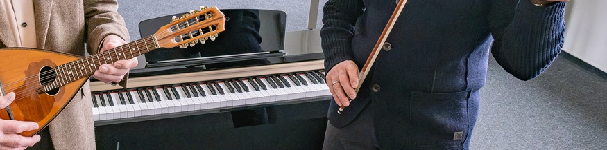 Zwei ältere Herren spielen ein Instrument in einem Klassenzimmer/Musikschule: Laute und Violine, dahinter ein Keyboard