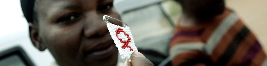 Eine Frau in Botswana hält eine Anstecknadel mit einem Aids Zeichen in die Kamera.