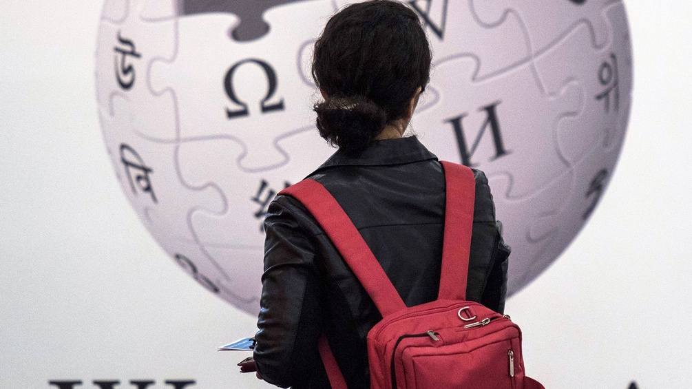 Frau mit Rucksack steht vor Wikipedia-Plakat