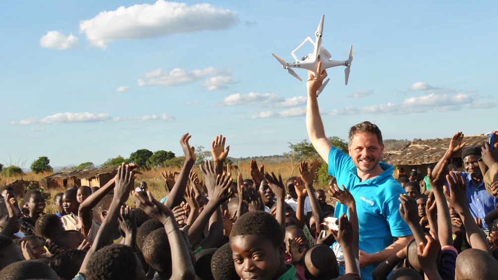 UNICEF-Mitarbeiter Michael Scheibenreif steht in Malawi inmitten einer Menge an Dorfbewohnern und hält eine Drohne in die Luft. 