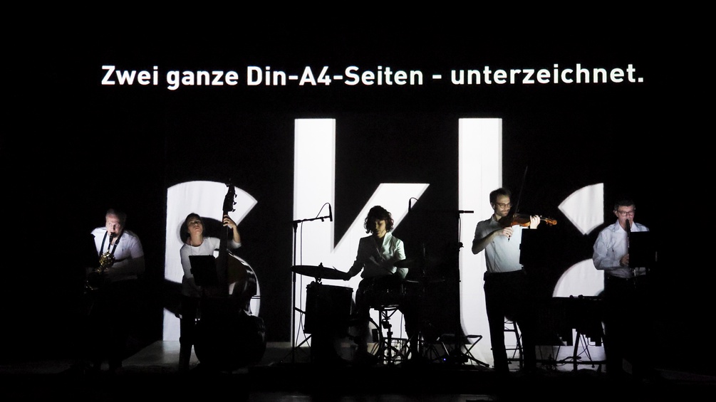 Musiker:innen spielen im Dunkeln, hinter ihnen weiße Schriftzeichen