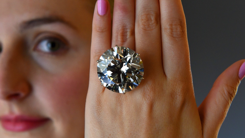 Eine Frau hält einen Ring mit einem riesen Diamant vor die Kamera.