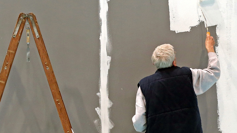 Älterer Mann streicht eine Wand weiß, daneben Leiter