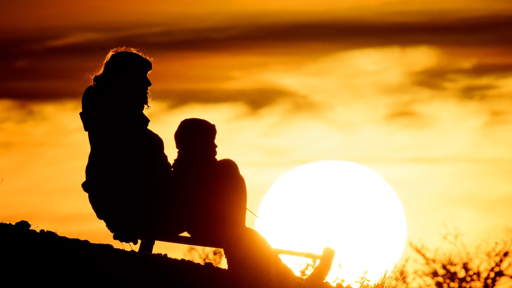 Eine Frau und ihr Sohn sitzen auf einer Bank und schauen auf den Sonnenuntergang