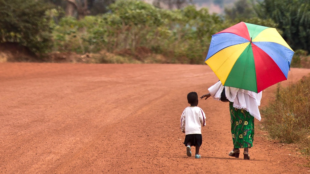 Frau und Kind samt Sonnenschirm gehen auf einer erdigen Straße in Ruanda