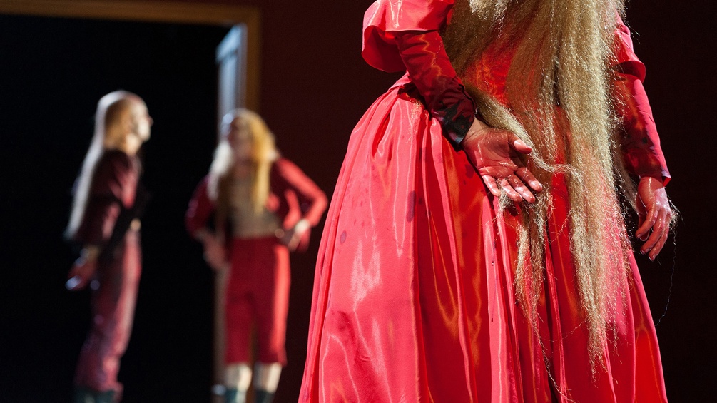 Person im roten Kleid miz blutigen Händen, im hintergrund zwei Figuren
