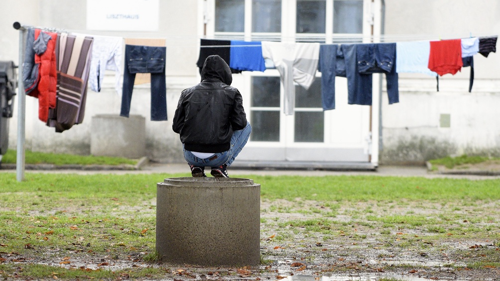 Mensch in Jacke hockt auf Betonbrunnenring, dahinter Wäsche auf einer Leine