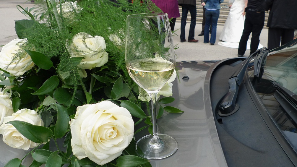 Weinglas, weiße Rosen, Brautpaar im Hintergrund