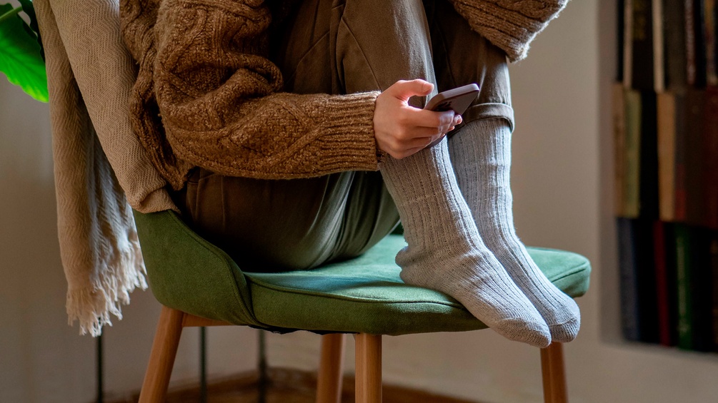 Junge Frau hockt mit angezogenen Beinen auf einem Sessel, in der Hand ein Smartphone