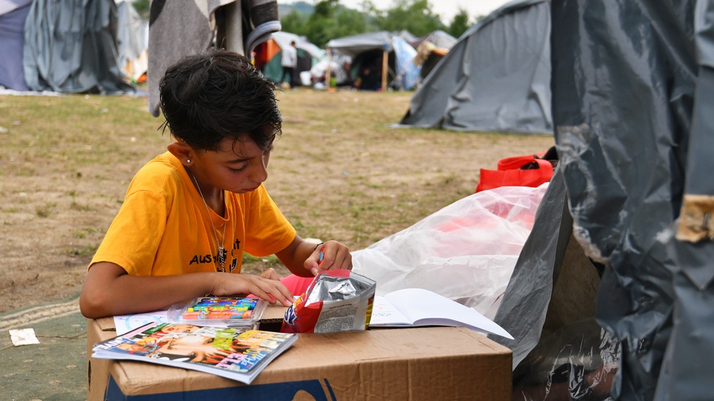Ein Flüchtlingskind zeichnet auf einem Tisch aus Karton, zwischen Zelten in Bosnien.
