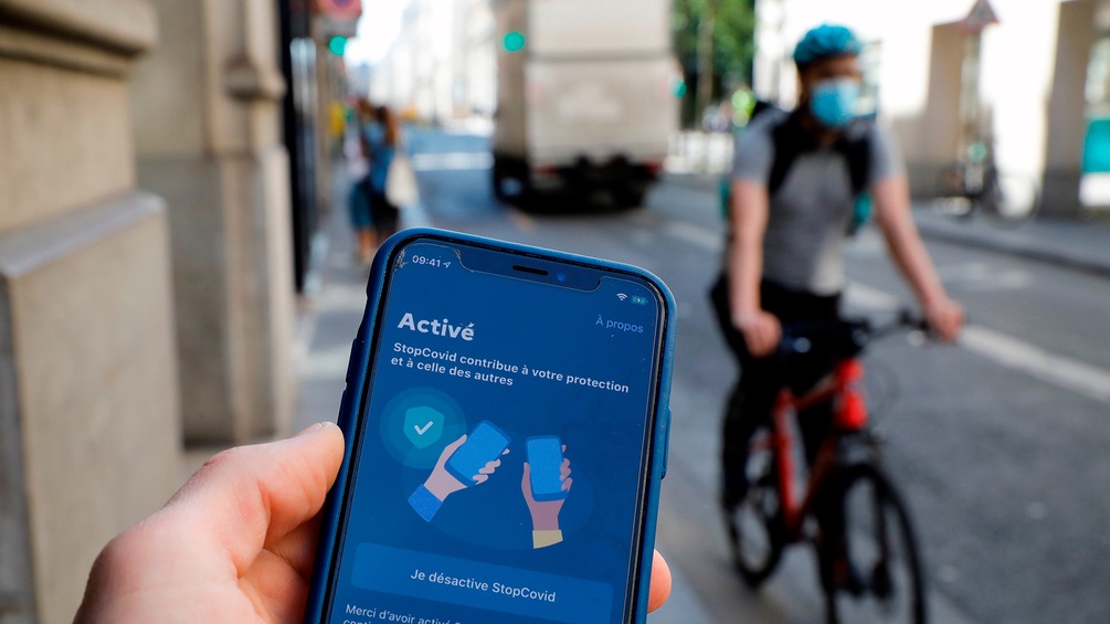 Mann hält in Frankreich ein Smartphone in Händen, in Hintergrundunschärfe ein Radfahrer mit Mundschutz