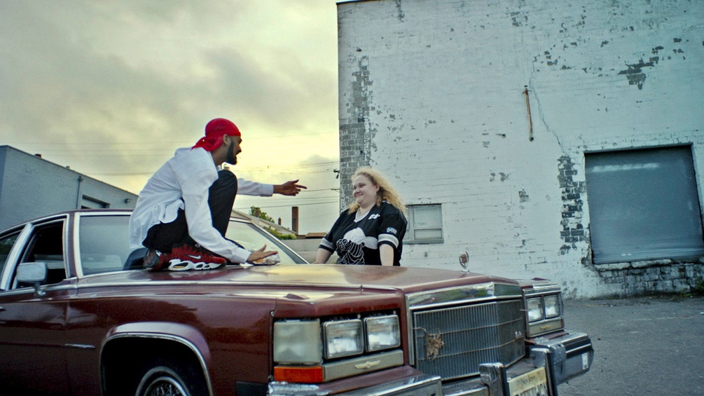 Rapper auf Motorhaube zeigt auf Frau mit blonden Haaren