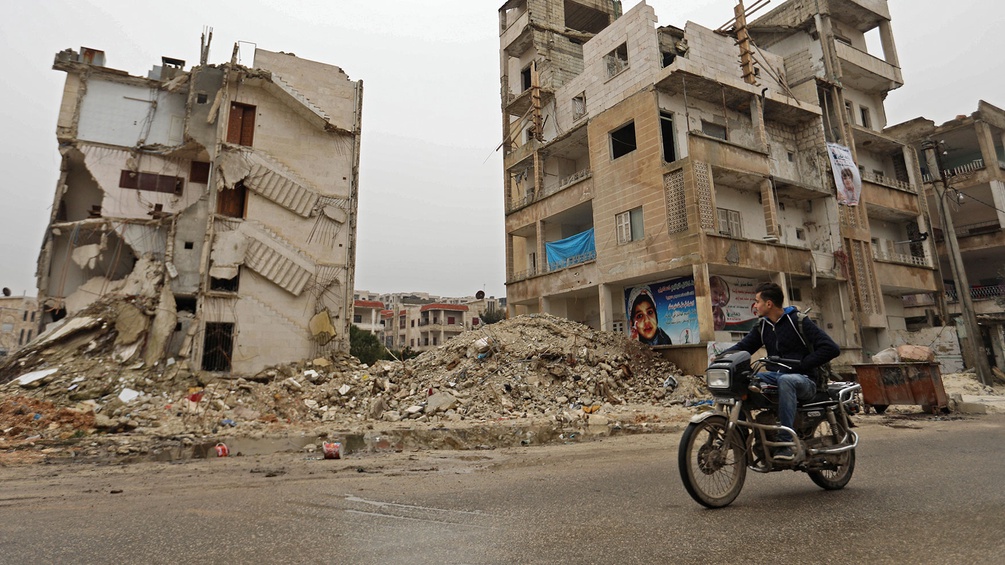 Mann auf einem Motorrad vor einer zerstörten Häuserzeile in Syrien