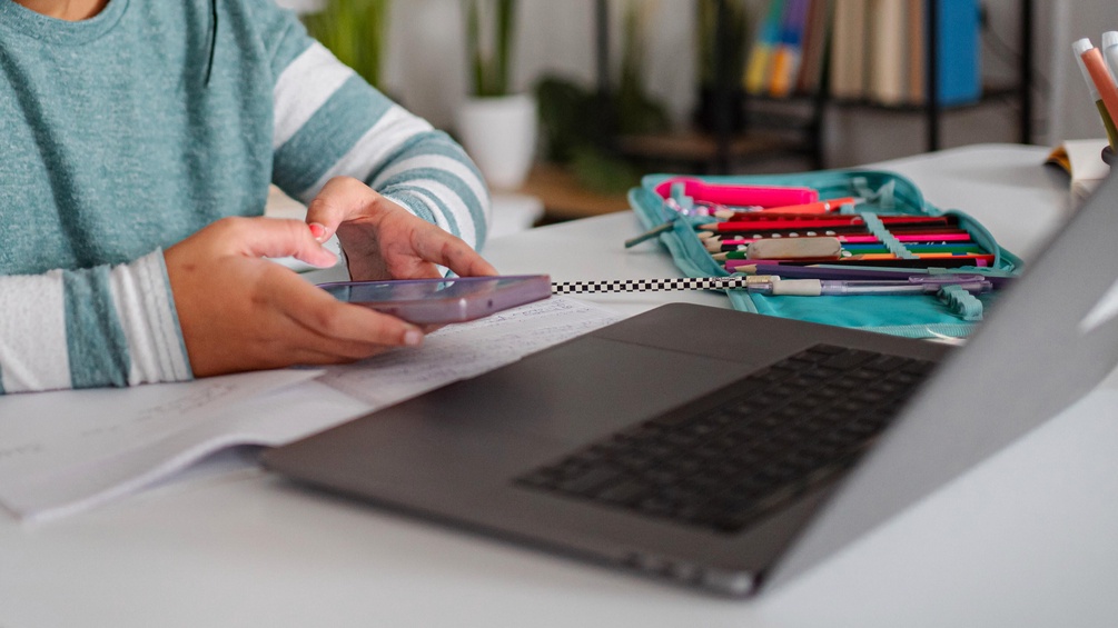 Kind hält Smartphone in Händen und sitzt an einem Tisch mit Laptop und Stiften