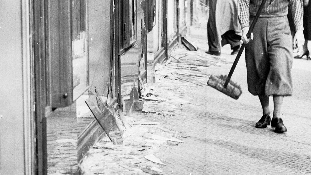 Ein junger Mann will am 10. November 1938 in Berlin mit einem Besen zerbrochene Scheiben zusammenkehren.