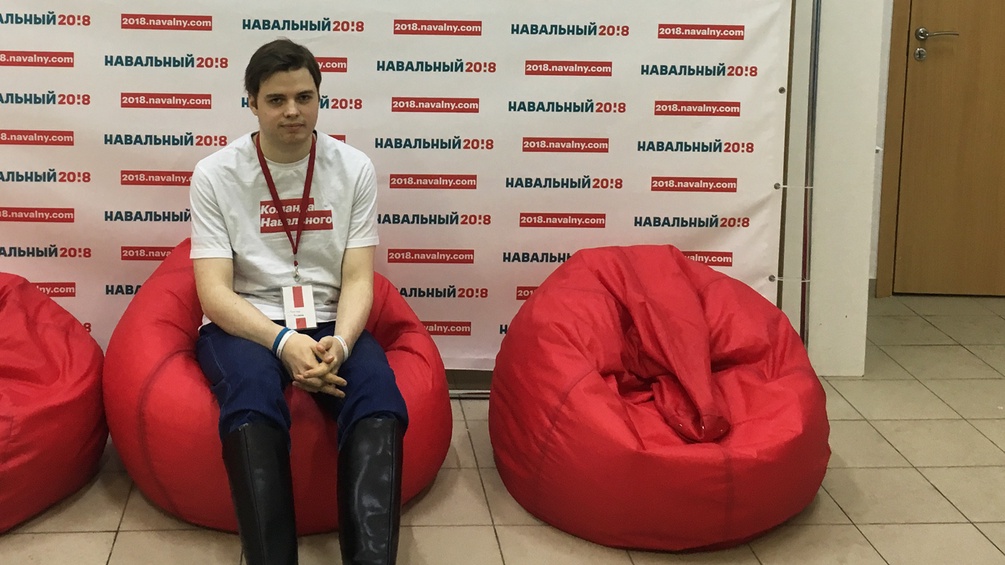 Ein junger Mann sitzt auf einem roten Sitzkissen. Im Hintergrund sieht man Wahlwerbung für Nawalny.