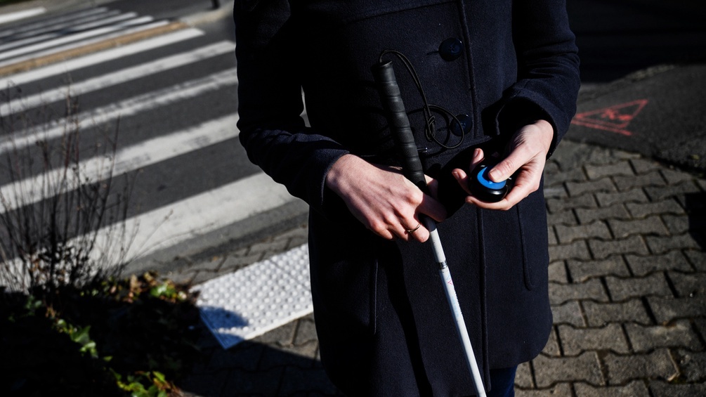 Torso einer Person mit Blindenstock in der einen Hand, einem elektronischen Gerät in der anderen.