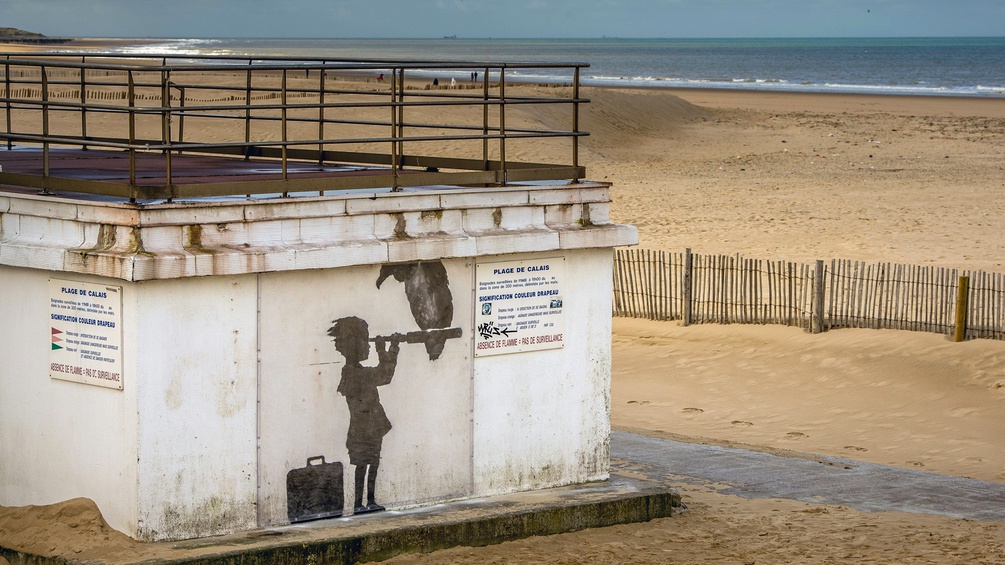 Strand von Calais 2015: Kind mit Koffer blickt durch ein Fernrohr, darauf sitzt ein Geier