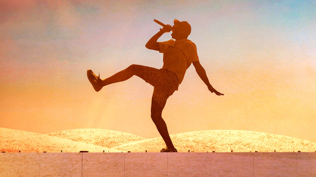 Ein Rapper tanzt mit Mikrophon auf einer Mauer, dahinter Sandhügel.