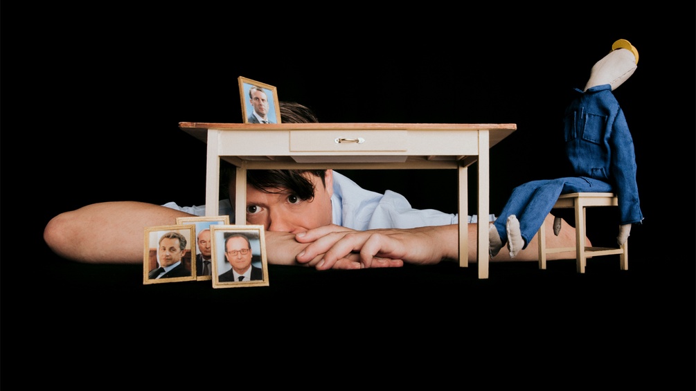 Das Plakat vom Stück "Wer hat meinen Vater umgebracht" - Ein junger Mann lehnt vor einem Puppentisch mit Fotos von französischen Politikern.
