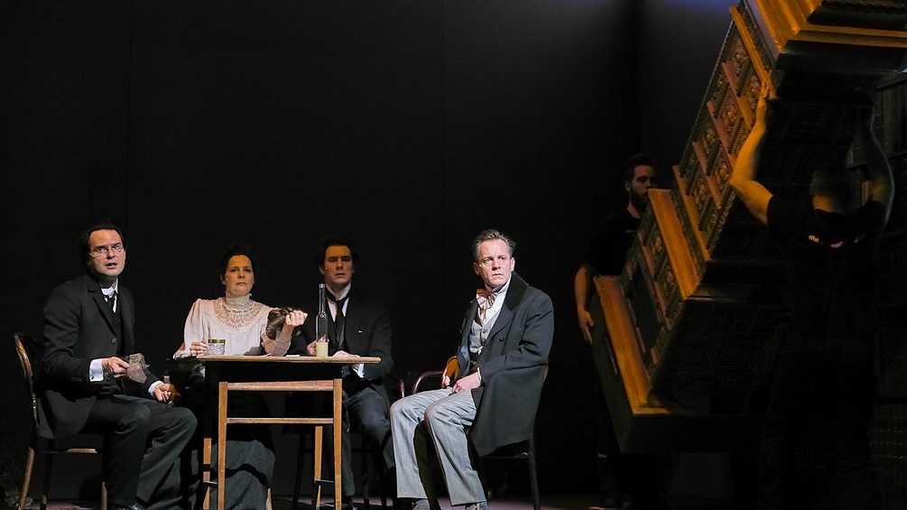 Szenenfoto: Drei Männer und eine Frau sitzen an einem Tisch, im Hintergrund wird ein Kachelofen fortgetragen