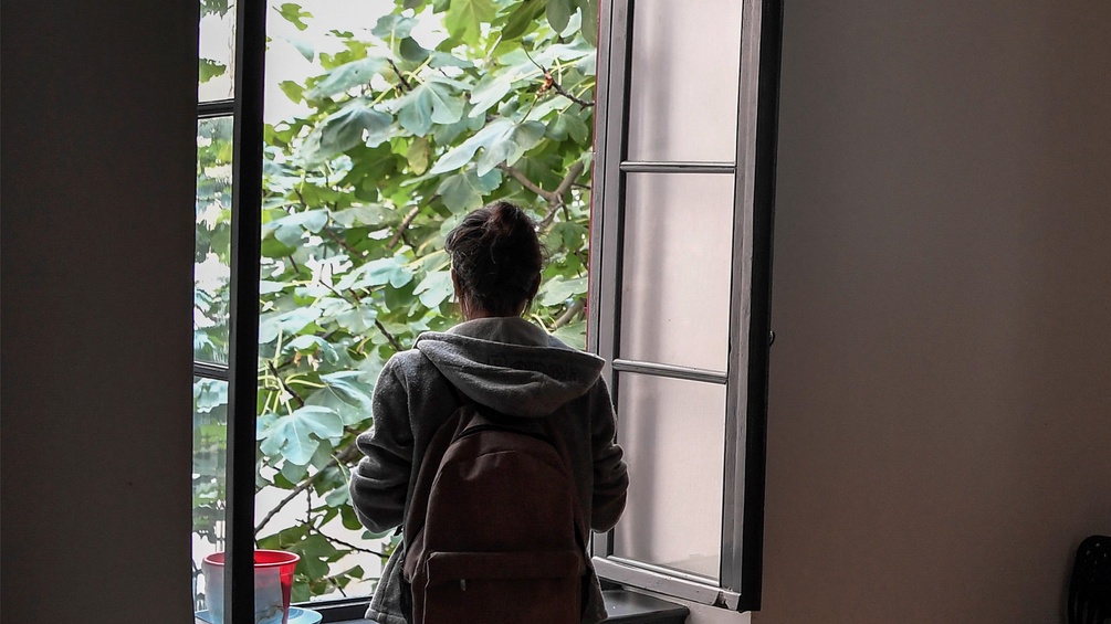 Eine Frau mit Rucksack steht vor dem Fenster und schaut ins Grüne hinaus.