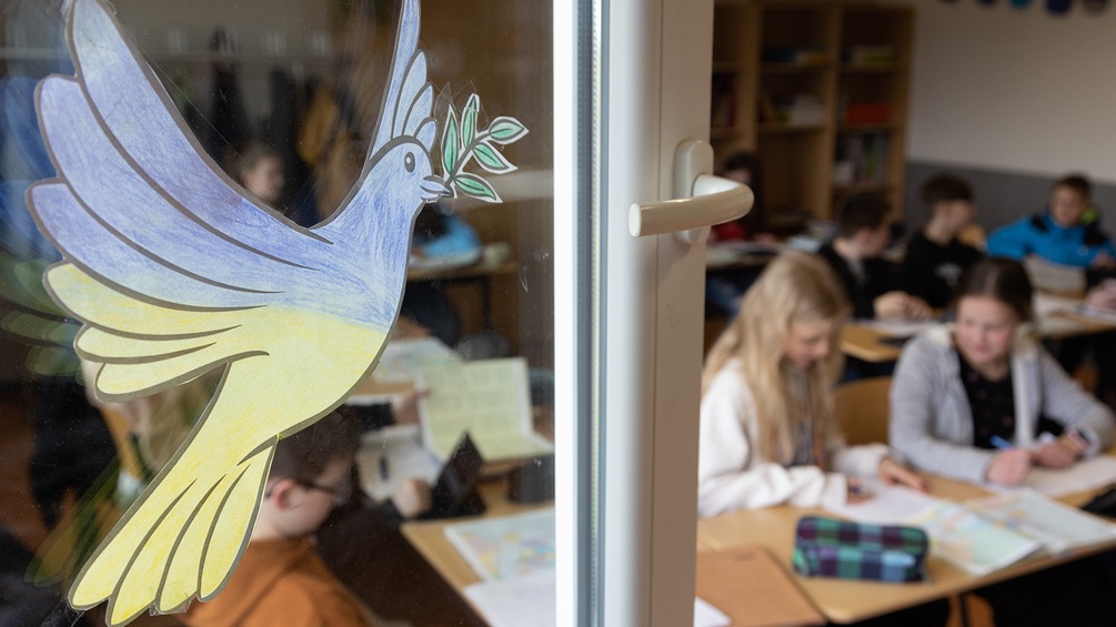 Eine Friedenstaube aus Papier, bemalt in blau und gelb, klebt auf dem Fenster eines Klassenzimmers.