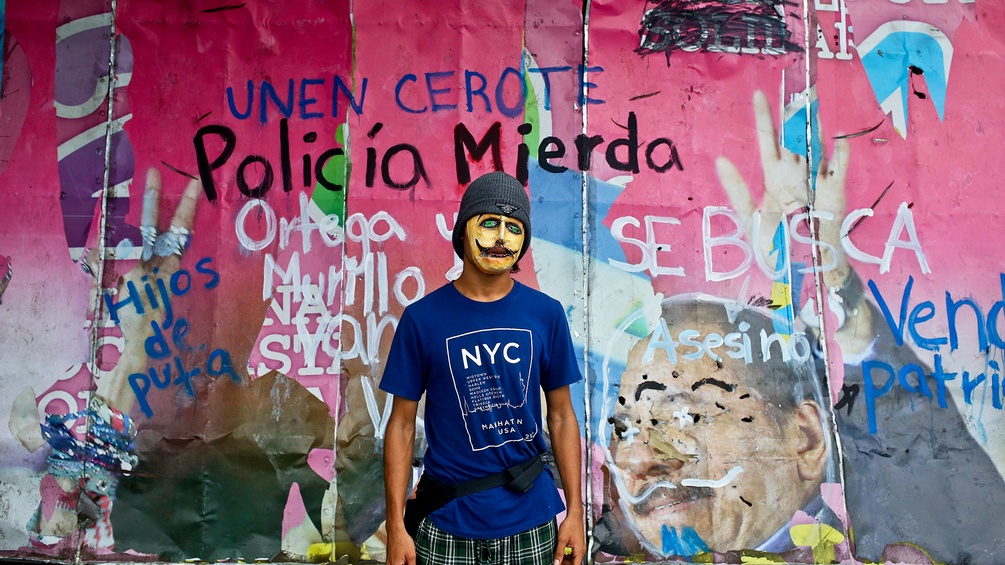 Ein Protestierender mit Maske in Nicaragua vor einem verunstalteten Plakat von Präsident Daniel Ortega 