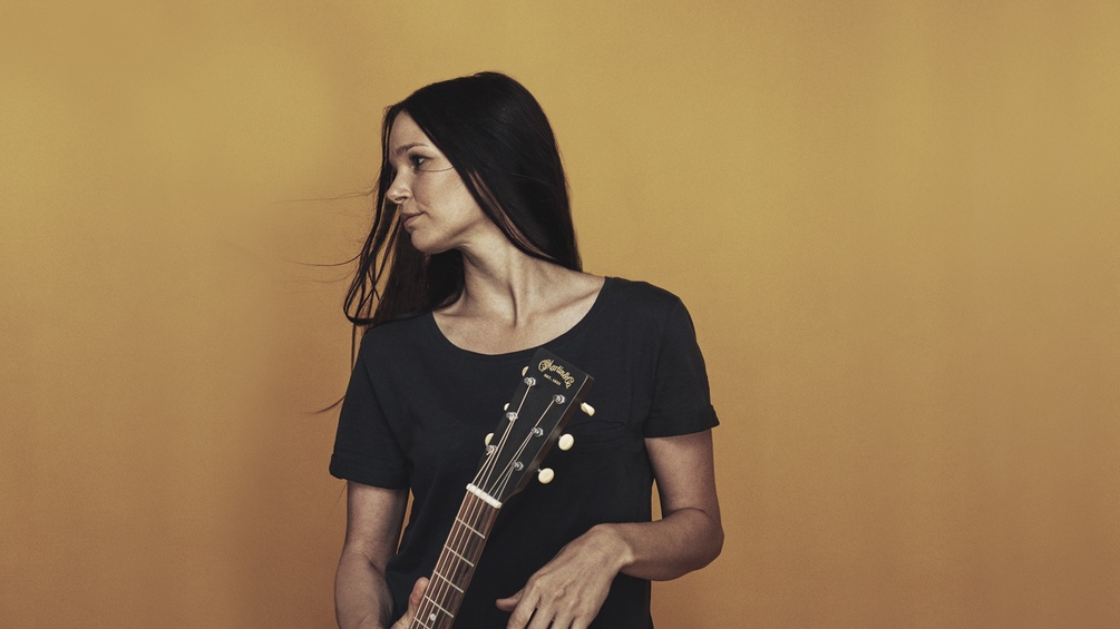 Susan Wolf mit Gitarre vor einem gelben Hintergrund