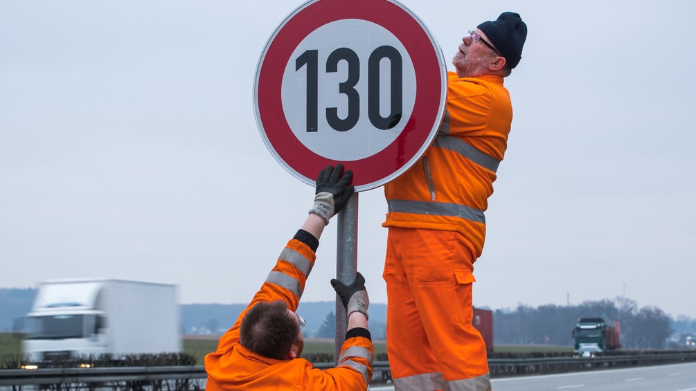 Zwei Männer montieren ein Schild auf der Autobahn mit der Ziffer 130.