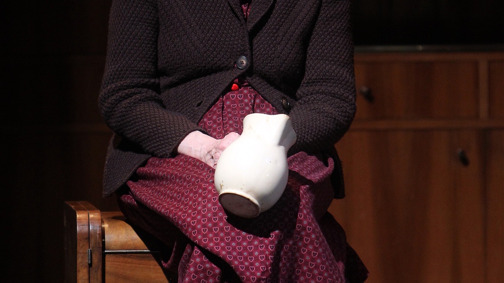 Eine sitzende ältere Frau mit Wasserkrug in der Hand.