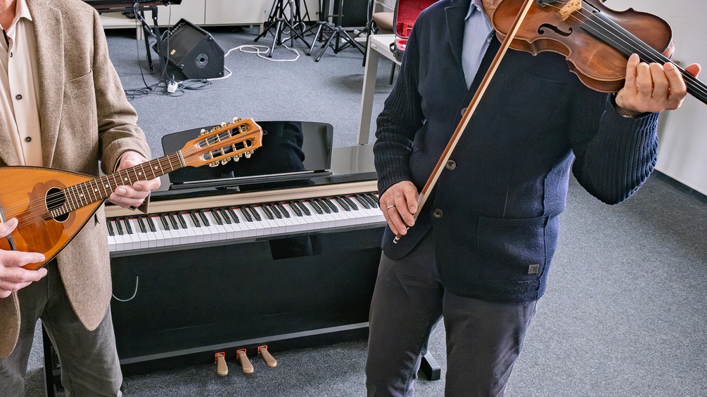 Zwei ältere Herren spielen ein Instrument in einem Klassenzimmer/Musikschule: Laute und Violine, dahinter ein Keyboard