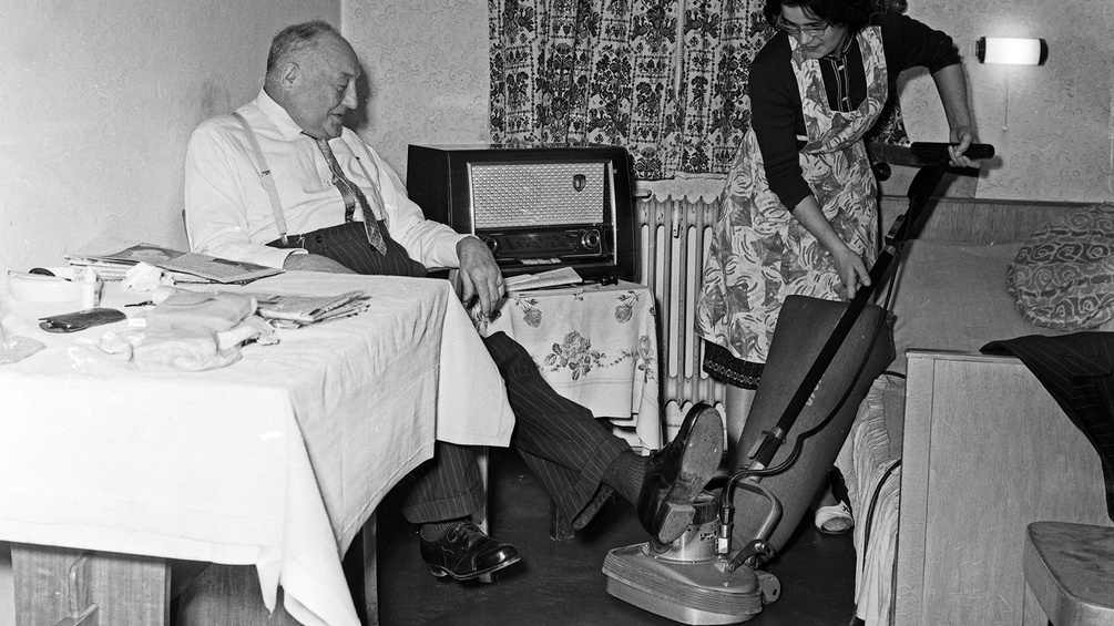 Deutschland, 1960er Jahre: Eine Frau saugt Staub, der Mann sitzt und hebt die Füße