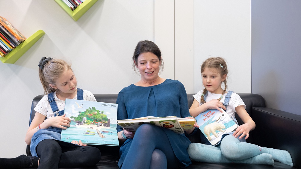 Begeisterung für das Buch: Die KiJuBuTAGE. Eine Frau mit zwei Mädchen sitzen auf einem Sofa mit Kinderbüchern.