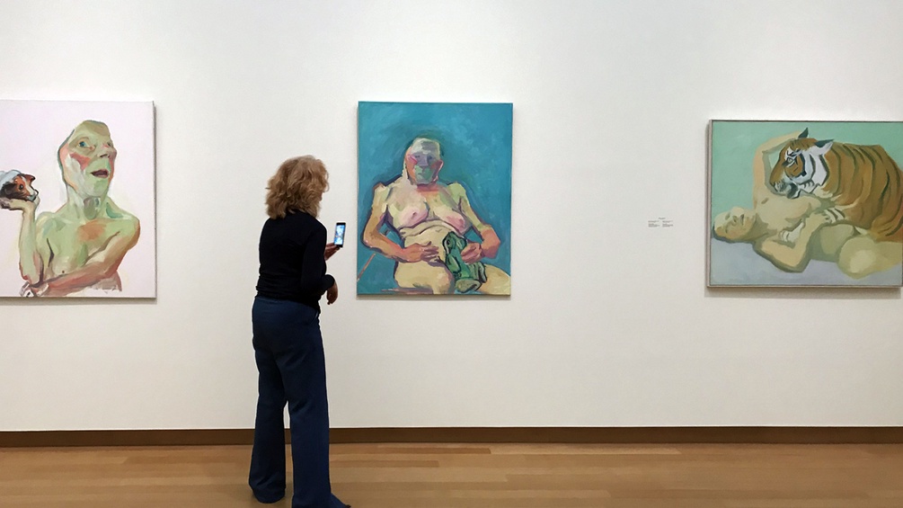 Eine Frau steht vor einem Maria Lassnig Werk und fotografiert es mit ihrem Smartphone