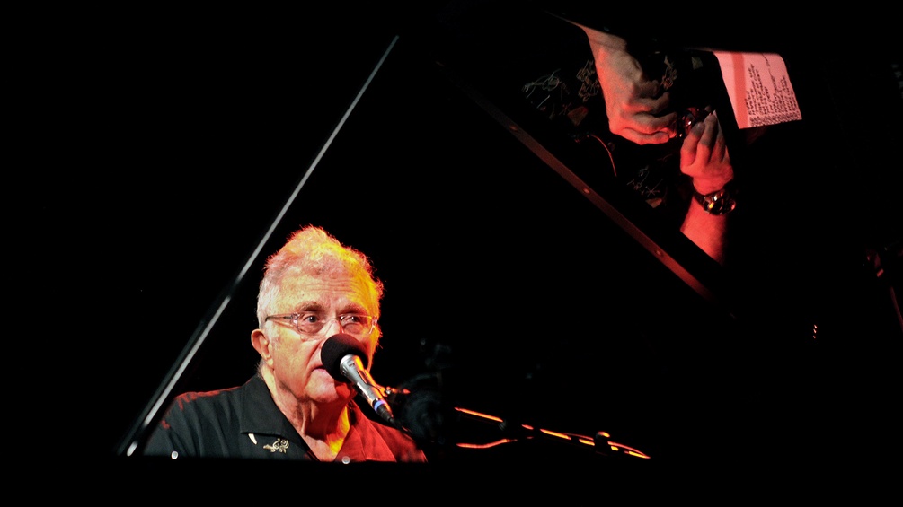 Randy Newman sitzt am Klavier. Das Mikrophon verdeckt sein halbes Gesicht.