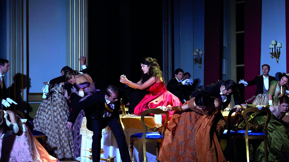 Szene aus "Teseo", Frau in einem Roten Kleid umgeben von anderen DarstellerInnen
