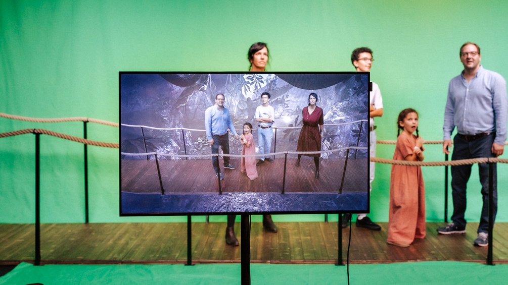 Eine Familie steht vor einem Greenscreen und hinter einem Bildschirm der sie scheinbar auf einer Brücke im Dschungel stehen lässt