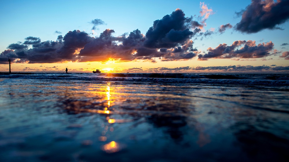 Norderney: Die Sonne geht hinter Wolken am Nordstrand der Insel unter und spiegelt sich dabei im Wasser der Nordsee.