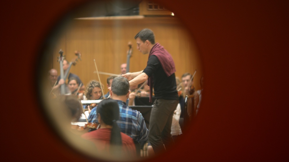 Blick durch ein rundes Fenster auf Dirigenten & Orchester
