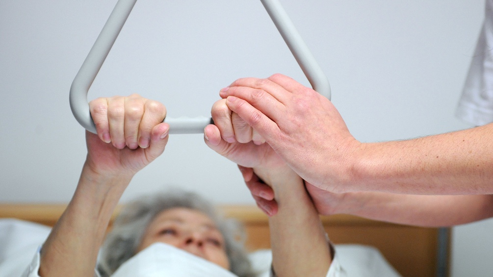 Eine Patientin hält sich an einer Triangel-Aufrichtungs-Hilfe fest und wird von einer Diplomkrankenschwester betreut 