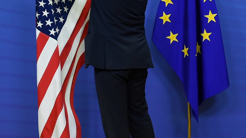 Ein Mann steht zwischen der Amerika und der EU-Flagge.